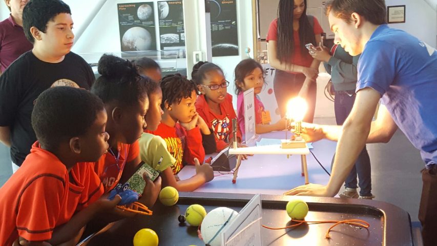 Students gathered at a table as a museum educator demonstrates a glowing light bulb