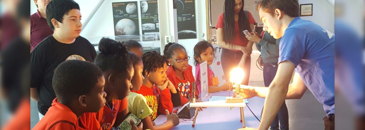 Students gathered at a table as a museum educator demonstrates a glowing light bulb